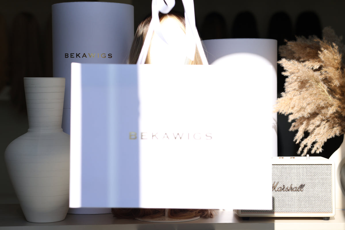 A white BEKAWIGS branded shopping bag in the foreground, with matching white cylindrical packaging and decorative home decor items including a vase, pampas grass, and a Marshall speaker in the background.